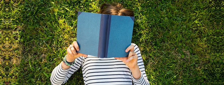 A person in a striped shirt laying in the grass with a blue, hardcover book blocking their face.